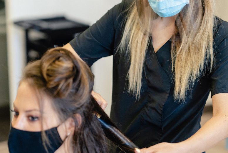 Hairdresser, protected by a mask, combing her clients hair with a hair iron in a salon