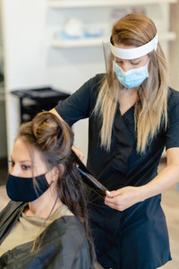 Hairdresser  protected by a mask  combing her clients hair with a hair iron in a salon
