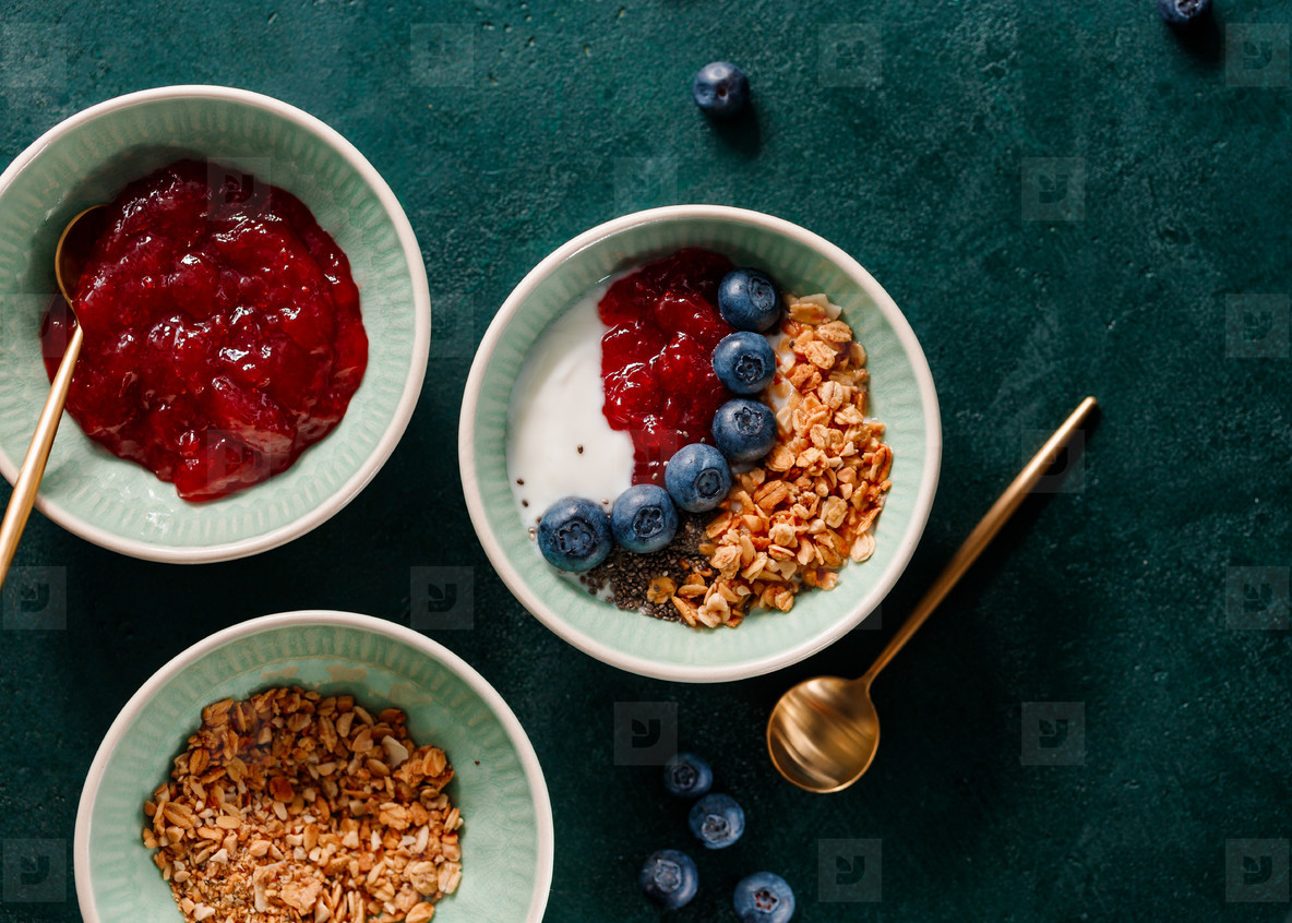 Top view of breakfast with granola yoghurt strawberry jam chia seeds and blueberry on a dark green table