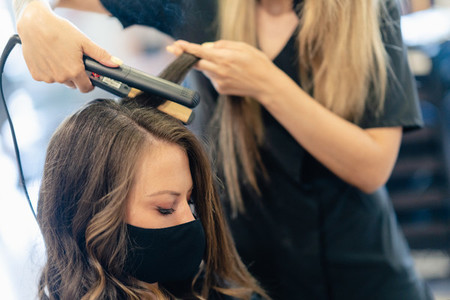 Hairdresser  protected by a mask  combing her clients hair with a hair iron in a salon