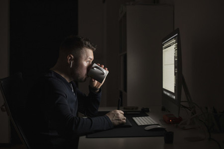 Man with earbuds drinking coffee and working late at computer in dark room