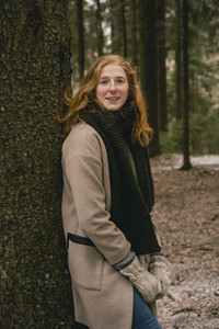 Portrait confident redhead woman in scarf and wool coat leaning against tree in woods