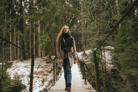 Woman in wool coat and scarf walking on footbridge in snowy woods