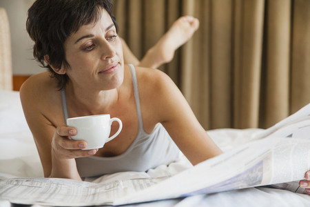 Woman relaxing on bed with coffee and reading newspaper