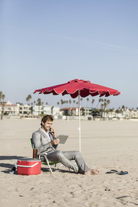 Barefoot businessman with smart phone and digital tablet working on beach