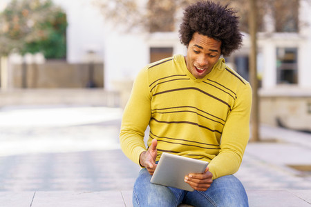 Black man using a digital tablet sitting on a bench outdoors