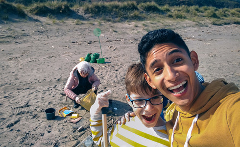 Young man and boy taking selfie while group of volunteers cleaning beach