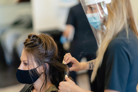 Hairdresser protected by a mask combing her clients hair with a hair iron in a salon