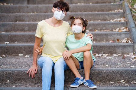 Middle aged mother and daughter sit on the street wearing masks