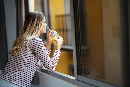 Young woman drinking a glass of natural orange juice  leaning out the window of her home