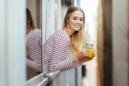 Smiling woman drinking a glass of natural orange juice  leaning out the window of her home