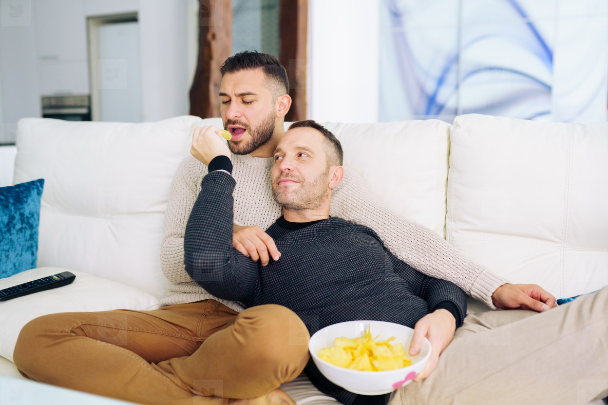 Gay couple sitting on the couch at home watching something on TV and having a snack