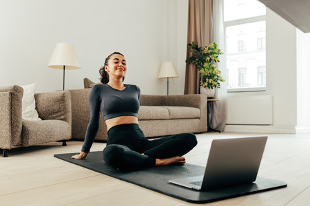 Smiling woman in sport clothes sitting on a mat in living room in front of a laptop
