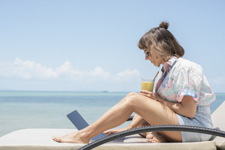 Woman with cocktail using laptop on sunny beach lounge chair