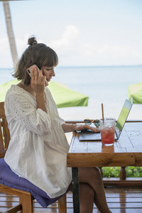 Woman with smart phone and laptop working at beach cafe table