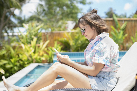 Woman using smart phone on sunny poolside lounge chair