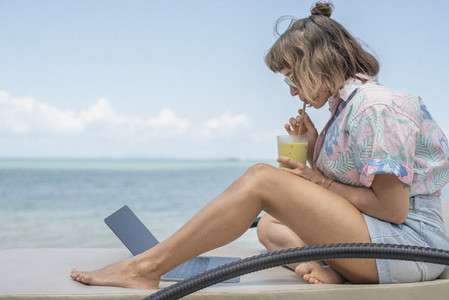 Woman sipping cocktail at laptop on beach lounge chair