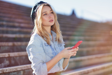 Blonde woman using a smartphone sitting on some city steps  wearing a denim shirt and black beret