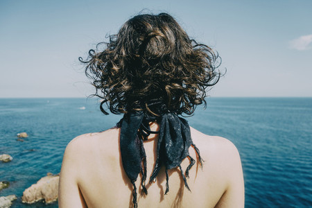 A girl with a kerchief on her neck and her back naked from behind looking at the horizon in the sea