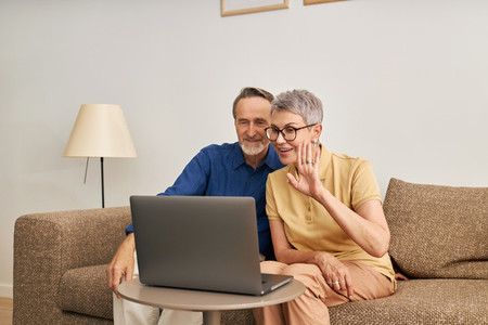 Cheerful senior couple using a laptop for video calling from the living room  Woman and man sitting