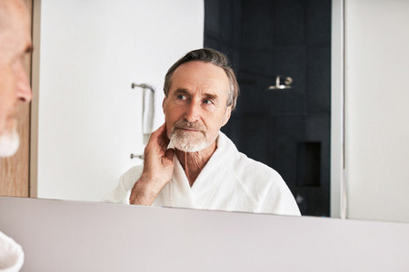Bearded mature man inspecting his skin in front of a mirror