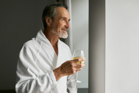 Smiling senior man with wineglass Side view of a bearded mature male wearing a bathrobe in a hotel room looking at window