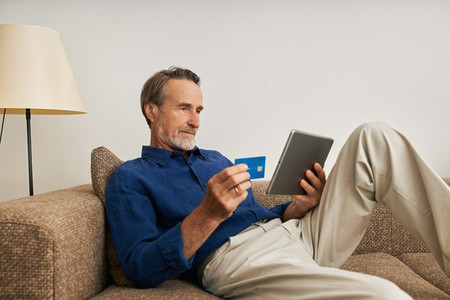 Handsome bearded man holding a digital tablet and credit card while sitting on a couch at home