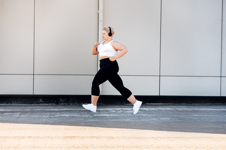 Side view of plus size woman wearing headphones running near a wall  Curvy female in sports clothes jogging outdoors