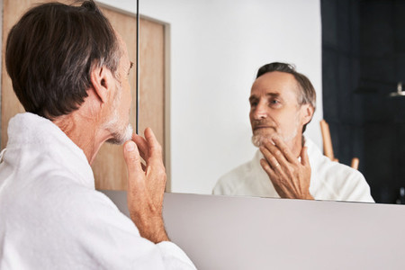Senior man touching his chin in bathroom in front of a mirror
