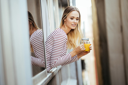 Young woman drinking a glass of natural orange juice  leaning out the window of her home