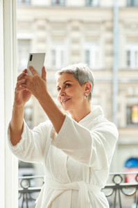 Smiling mature woman in bathrobe taking a selfie while standing on a balcony