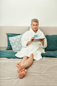 Mature woman in bathrobe lying on a bed holding a digital tablet and credit card  Senior female ordering from a hotel room