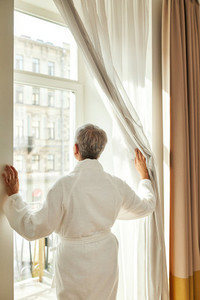 Back view of mature woman in bathrobe pulls the curtain and looks out while standing in hotel room