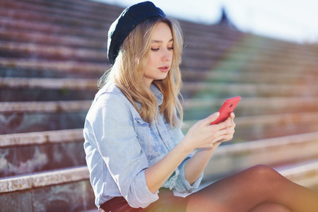 Blonde woman using a smartphone sitting on some city steps  wearing a denim shirt and black beret