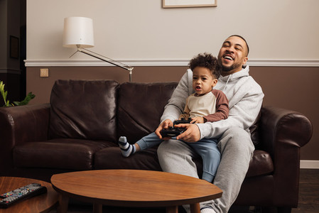 Father and son playing a video game together at home  Young man and little boy sitting on sofa holding joysticks and having fun