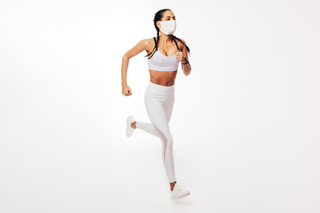 Young fit woman in face mask running on white background in studio