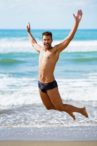Young man with beautiful body in swimwear jumping on a tropical beach