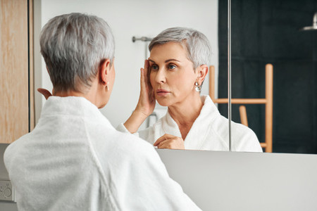 Senior female touching her face in a bathroom Mature woman inspecting face in the morning
