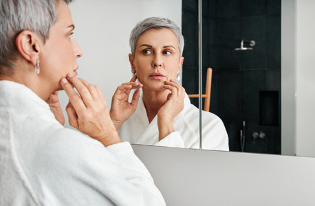 Mature woman with short grey hair wearing bathrobe standing in front of a bathroom mirror