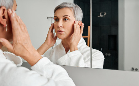 Mature woman in bathrobe looking herself in the bathroom mirror