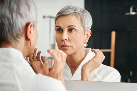 Senior woman with moisturizer on her face looking at mirror in bathroom