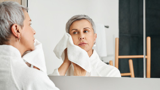Beautiful mature woman doing morning routine in bathroom wiping the face