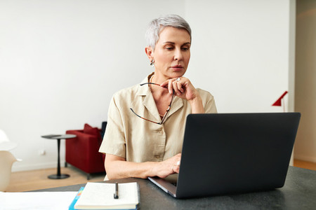 Mature adult female holding glasses typing on a laptop at home