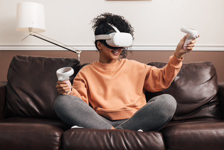 Woman with joysticks and VR goggles playing video games on a sofa