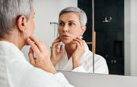 Mature woman with short grey hair touching her face in front of a bathroom mirror