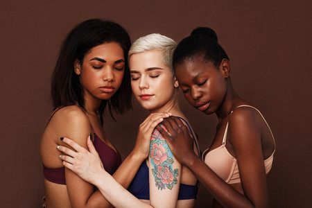 Three diverse women in bras standing with closed eyes over brown background