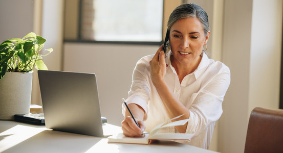 Businesswoman talking on cell phone and taking notes in office