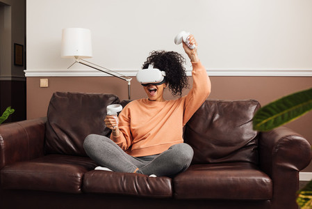 Excited female gamer using virtual reality headset while sitting in room on a couch