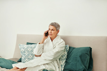Mature woman in a bathrobe talking on mobile phone while lying on a bed in hotel room