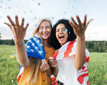 Two laughing female friends throwing confetti in the air while standing on a field with USA flag
